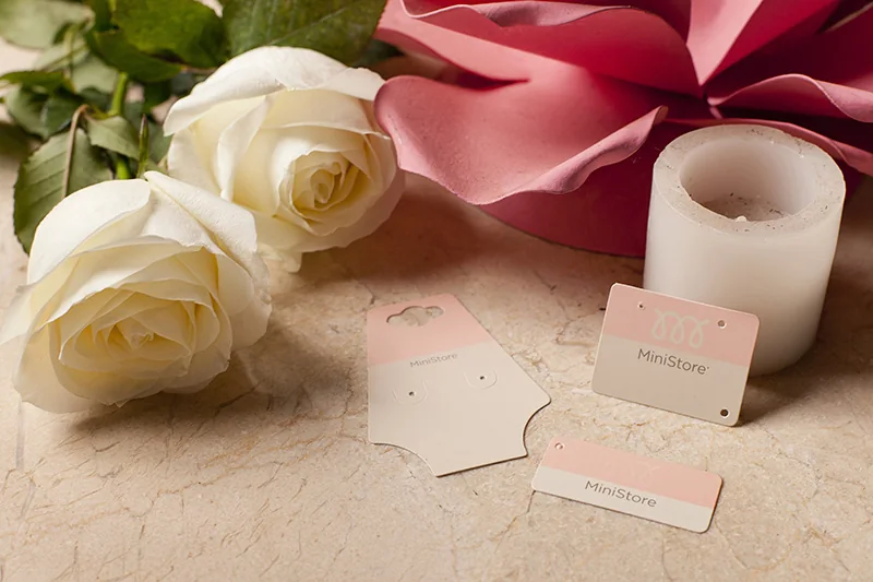 Tags de papel em rosa e off-white, dispostas com rosas brancas, laço de cetim e vela